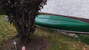 Un bateau vert, recouvert d’une bâche, partiellement dissimulé par un arbre, avec un mur blanc à texture visible en arrière-plan.