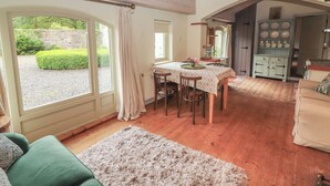 A cozy room with a dining table, chairs, a green sofa, a fluffy rug, and a view of the garden through large windows. - The Coach House Cottage (Dromineer)