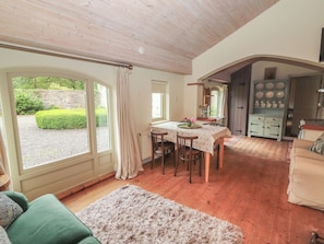 A cozy room with a dining table, chairs, a green sofa, a fluffy rug, and a view of the garden through large windows. - The Coach House Cottage (Dromineer)