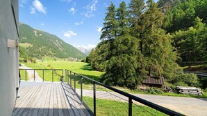 Une terrasse en bois avec une rambarde en métal, offrant une vue sur un paysage verdoyant, avec des arbres et une montagne à l’horizon.