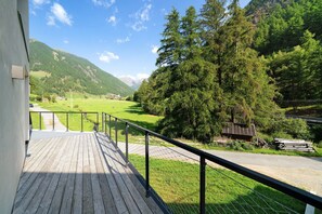 A wooden deck with a metal railing, overlooking a green landscape with trees and a mountain in the distance.