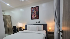 A hotel room with a bed, a desk, a chair, a nightstand, a lamp, a painting on the wall, and a door. - Devon's Stylish 1BR Apartment + Pool, Apo Resettlement Abuja (Abuja)