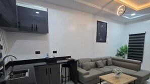 A modern living room with a sofa, a coffee table, a kitchen area with a sink, and a ceiling light fixture. - Devon's Stylish 1BR Apartment + Pool, Apo Resettlement Abuja (Abuja)