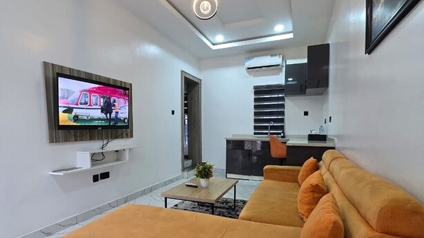A modern living room with a flat-screen TV, a sofa, a coffee table, and a kitchen area with cabinets and an air conditioning unit. - Devon's Stylish 1BR Apartment + Pool, Apo Resettlement Abuja (Abuja)