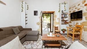 A living room with a sofa, a wooden table, a TV mounted on a stone wall, and a bookshelf. - Xanthippi s Traditional House in Pemonia (Apokoronas)