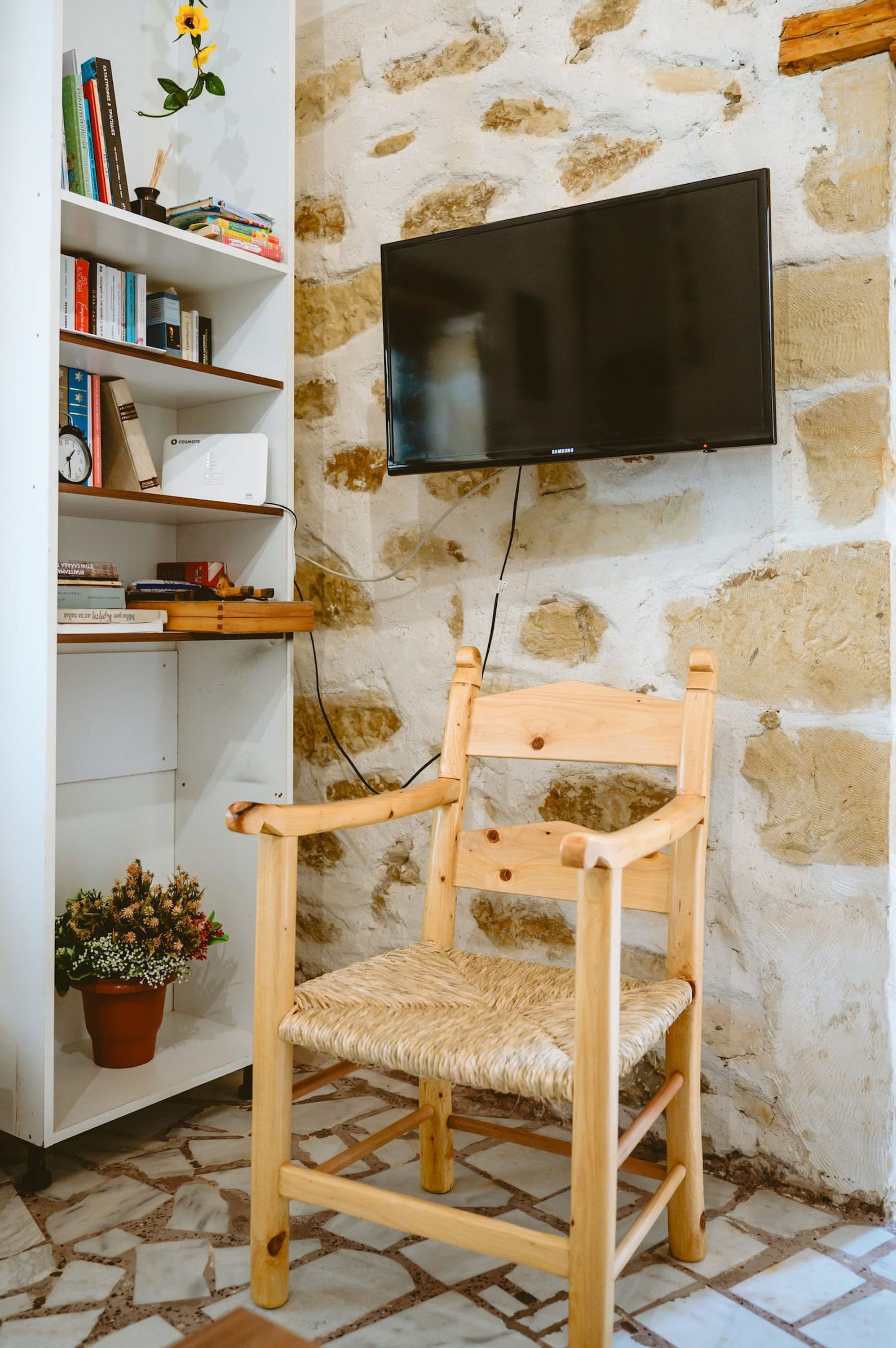 Een houten stoel met een geweven zitvlak, een witte boekenkast met boeken en decoratieve objecten, en een televisie bevestigd aan een stenen muur.