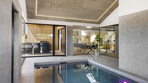 Un espace piscine intérieur moderne avec des murs en verre, un coin salon avec des chaises et une table, et un plafond doté d’un motif lumineux géométrique.