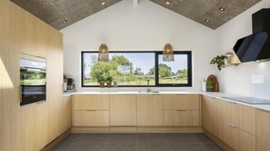 A modern kitchen with wooden cabinets, a built-in oven, a large window with a view of greenery, and pendant lighting. - VillaVilla 975 - Hasmark Strand, Fyn og øer (Otterup)