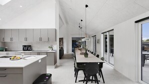 Eine moderne Küche mit Kochinsel, ein Essbereich mit langem Tisch und Stühlen sowie ein Blick durch eine Glastür auf einen Sitzbereich im Freien.
