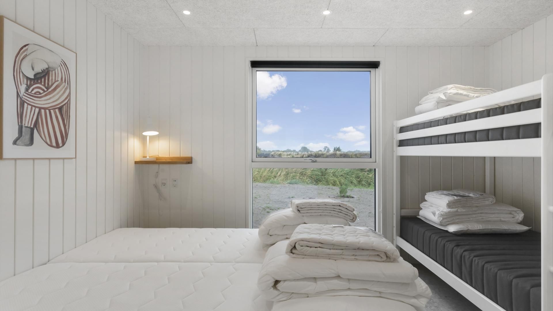 A bedroom with a bunk bed, a bedside table with a lamp, a window with a view of the outdoors, and a painting on the wall.