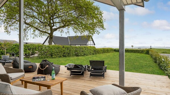 Ett takförsett utomhusområde med sittplatser, möbler i rotting, ett träbord och utsikt över ett hus och havet.