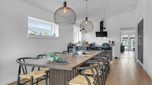 Cozinha moderna com uma mesa de jantar comprida, cadeiras, luminárias pendentes e uma TV na parede.