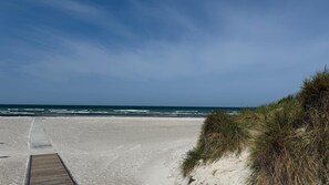Ein sandiger Strand mit einer hölzernen Promenade, die zum Wasser führt, und Dünen rechts.