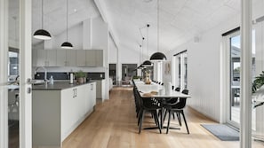 Una cocina moderna con una isla central, un comedor con una mesa larga y sillas, y vistas al exterior a través de amplios ventanales.