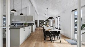 Eine moderne Küche mit einer zentralen Kochinsel, einem Essbereich mit einem langen Tisch und Stühlen sowie Blick ins Freie durch große Fenster.