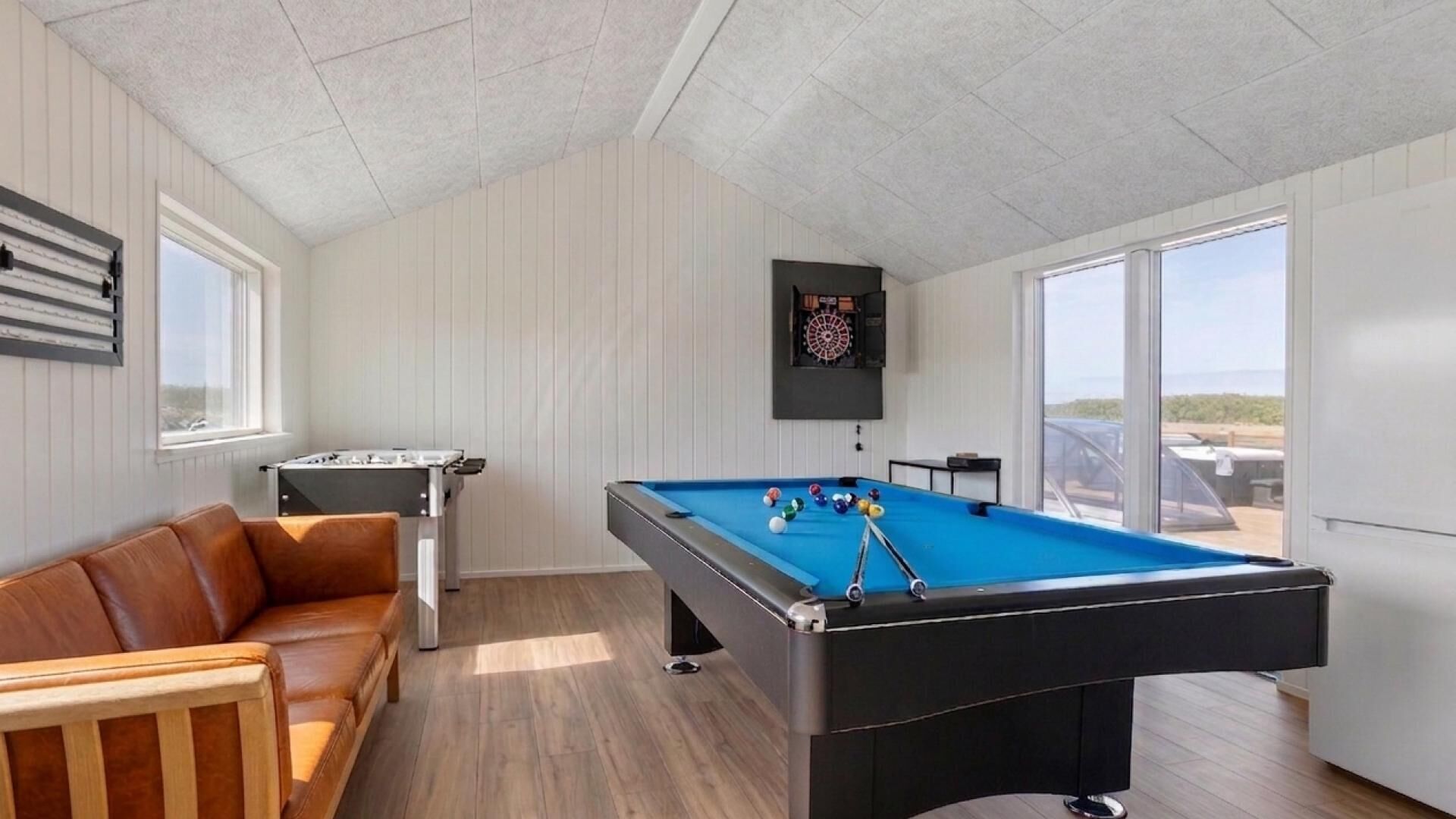 Une table de billard avec un jeu de fléchettes, une table de baby-foot et un canapé en cuir dans une pièce au plancher en bois et aux murs blancs.