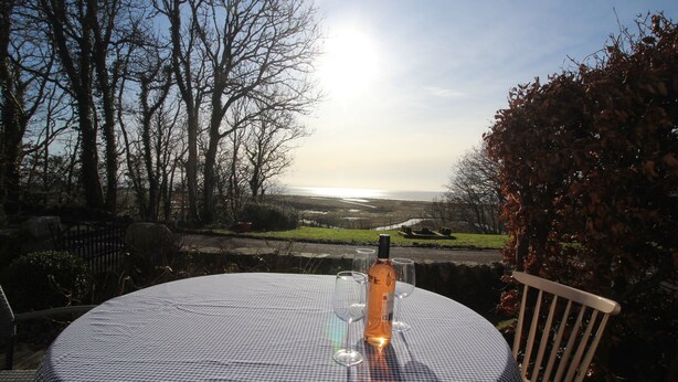 A table with a bottle and glasses, overlooking a landscape with trees and a body of water. - Rustic cottage with sea views (Southwick)