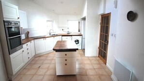 A kitchen with white cabinets, a wooden countertop, a built-in oven, a washing machine, and a window. - Rustic cottage with sea views (Southwick)