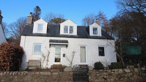 A white two-story house with a stone wall in front, a wooden bench, and a gate. - Rustic cottage with sea views (Southwick)