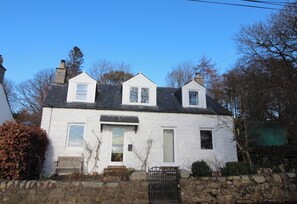 A white two-story house with a stone wall in front, a wooden bench, and a gate. - Rustic cottage with sea views (Southwick)
