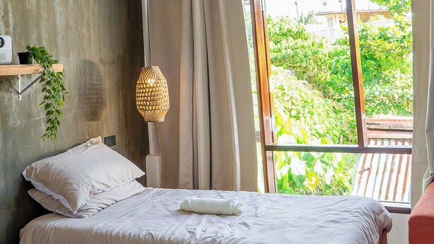 Une chambre à coucher avec un lit, une lampe suspendue, une fenêtre donnant sur un paysage verdoyant et une plante posée sur une étagère.