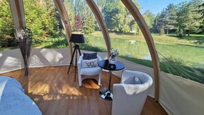 Habitación con tienda de campaña y vista a un lago, una lámpara con trípode, una silla blanca y una mesa pequeña con un arreglo floral.