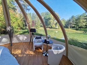Une pièce aménagée comme une tente, avec vue sur un lac, un lampadaire sur trépied, une chaise blanche et une petite table agrémentée d’un arrangement floral.