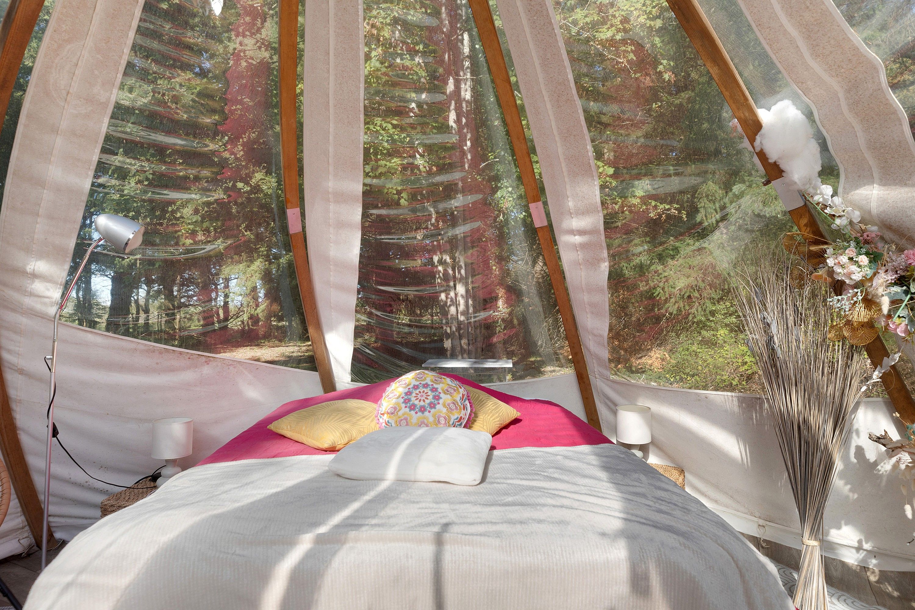 A tent-like room with a bed, a lamp, and a view of a forest.