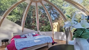 Una habitación acogedora con una cama, un taburete pequeño y un ventanal que da a un bosque.