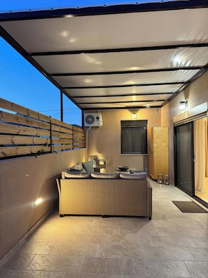 Eine Außenpatio mit einem Rattan-Sofa, einem Tisch, Stühlen, einer wandmontierten Klimaanlage und einer Glastür.