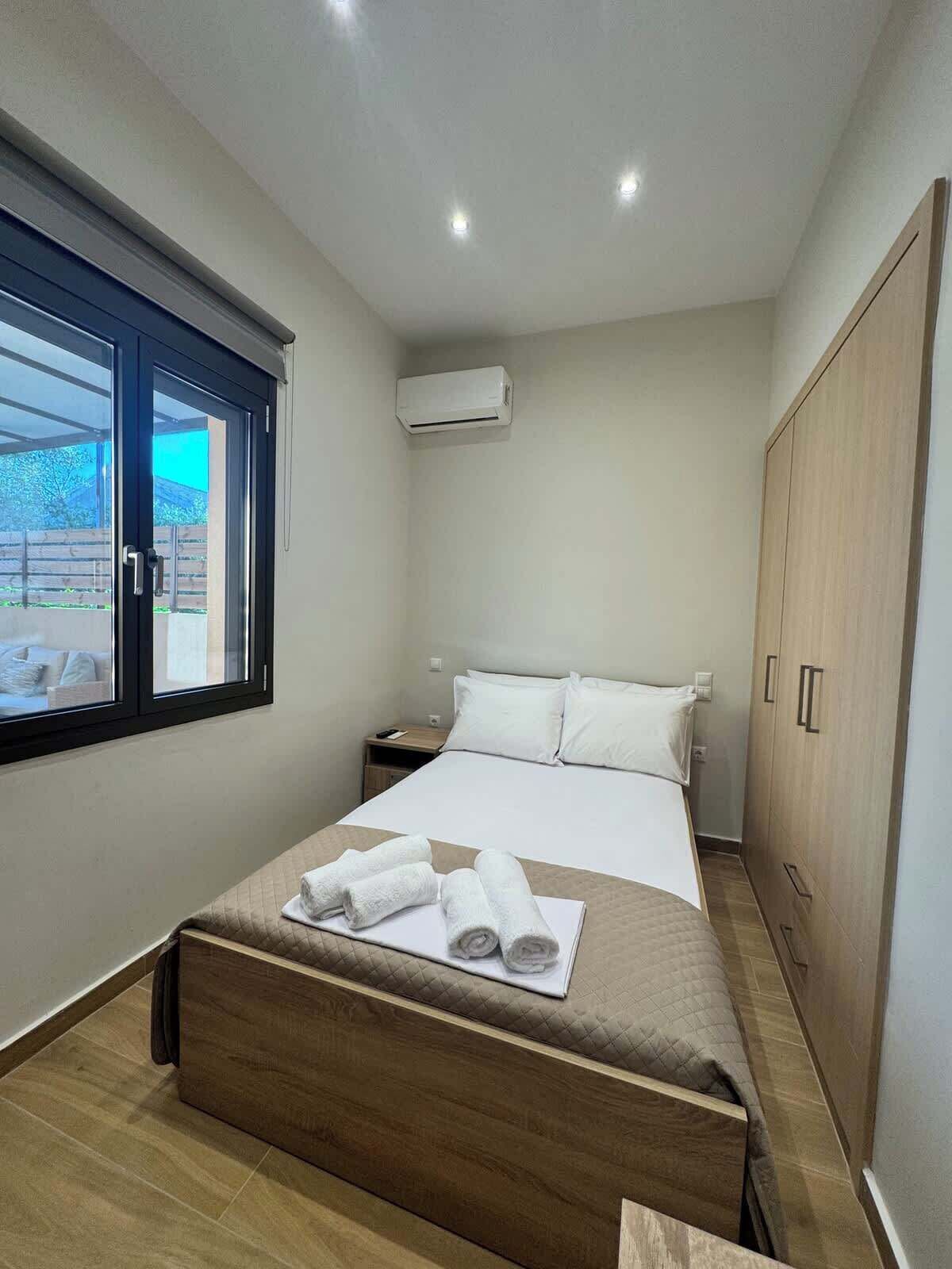 A bedroom with a bed, a window, an air conditioning unit, a wardrobe, and a bedside table.