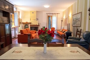 Ein Wohnzimmer mit Kamin, orangefarbenen und blauen Sofas, einer weißen Vase mit Blumen sowie einem Esstisch mit Platzdeckchen.