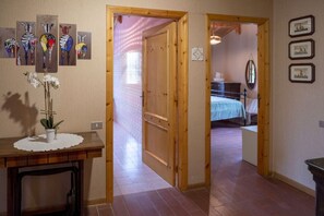 Ein Flur mit Holztüren, die zu einem Schlafzimmer mit Bett und Kommode führen. An den Wänden hängen Bilder, und ein kleiner Tisch trägt eine Pflanze.