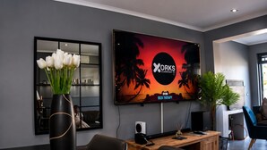 Ein moderner Arbeitsraum mit einem großen Bildschirm, der einen tropischen Sonnenuntergang zeigt, einem Schreibtisch mit einem Computer und einer Vase mit weißen Blumen.