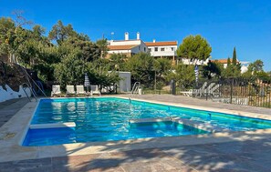 A swimming pool with a stone deck, surrounded by trees and a white building in the background. - Beautiful home in Laroya with outdoor swimming pool (Laroya)