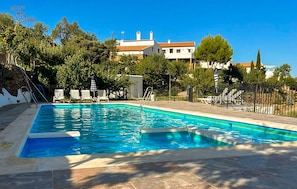 Ein rechteckiger Swimmingpool mit einer gefliesten Liegefläche, umgeben von Bäumen und einem weißen Gebäude mit rotem Dach im Hintergrund.