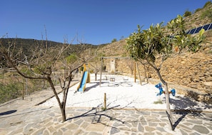 Ein Spielplatz mit Rutsche und Schaukeln, umgeben von Steinmauern und wenig Bepflanzung.