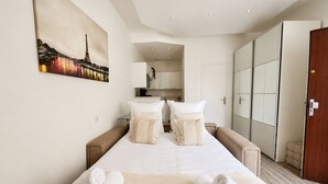 A hotel room with a bed, pillows, a painting of a cityscape, a kitchenette, and a wardrobe. - Luxueux Appartement au 16e  (Paris)