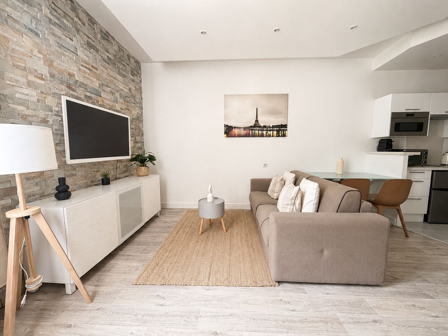 A modern living room with a stone wall, a flat-screen TV, a sofa, a coffee table, and a kitchen area with cabinets and appliances.