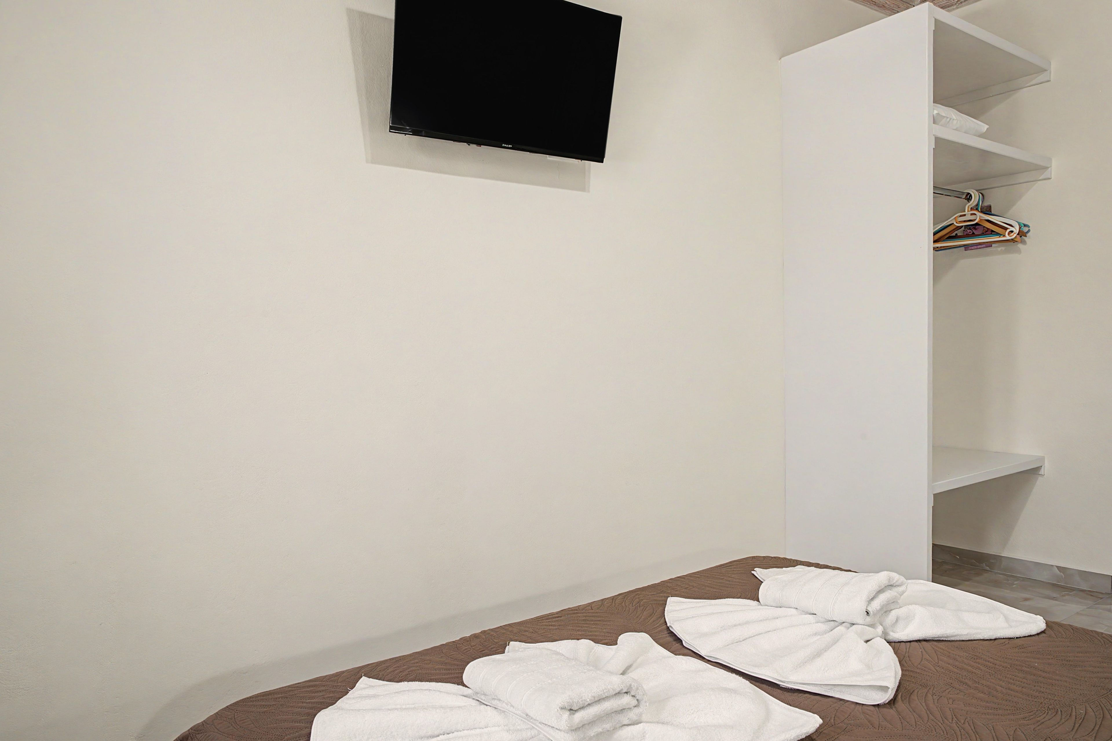 Une chambre avec un lit, un téléviseur à écran plat fixé au mur et une armoire blanche contenant des vêtements suspendus.