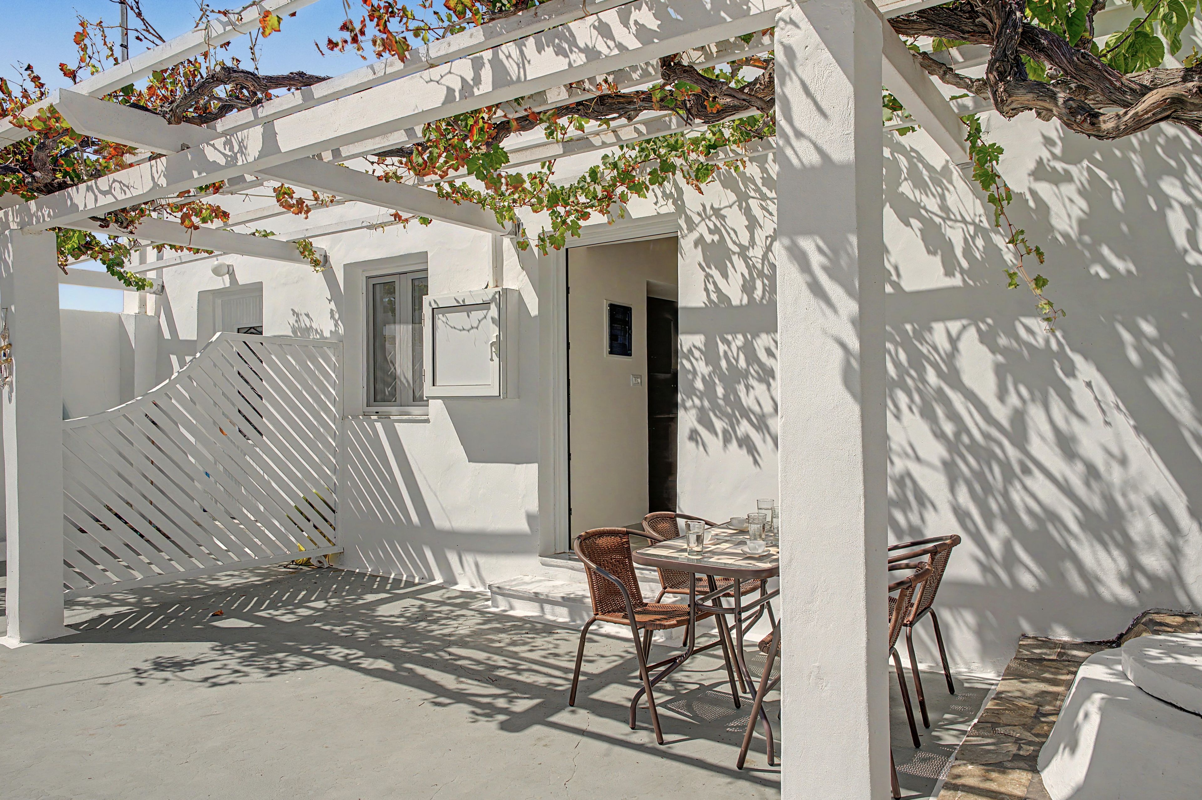 Een overdekt terras met een tafel en stoelen, een witte pergola en een gebouw met een raam en een deur.