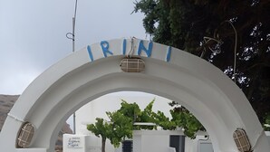 Una entrada arqueada blanca con el nombre 'IRINI' encima, un letrero de 'IRINI STUDIOS' a la izquierda, y una mesa con sillas al fondo.