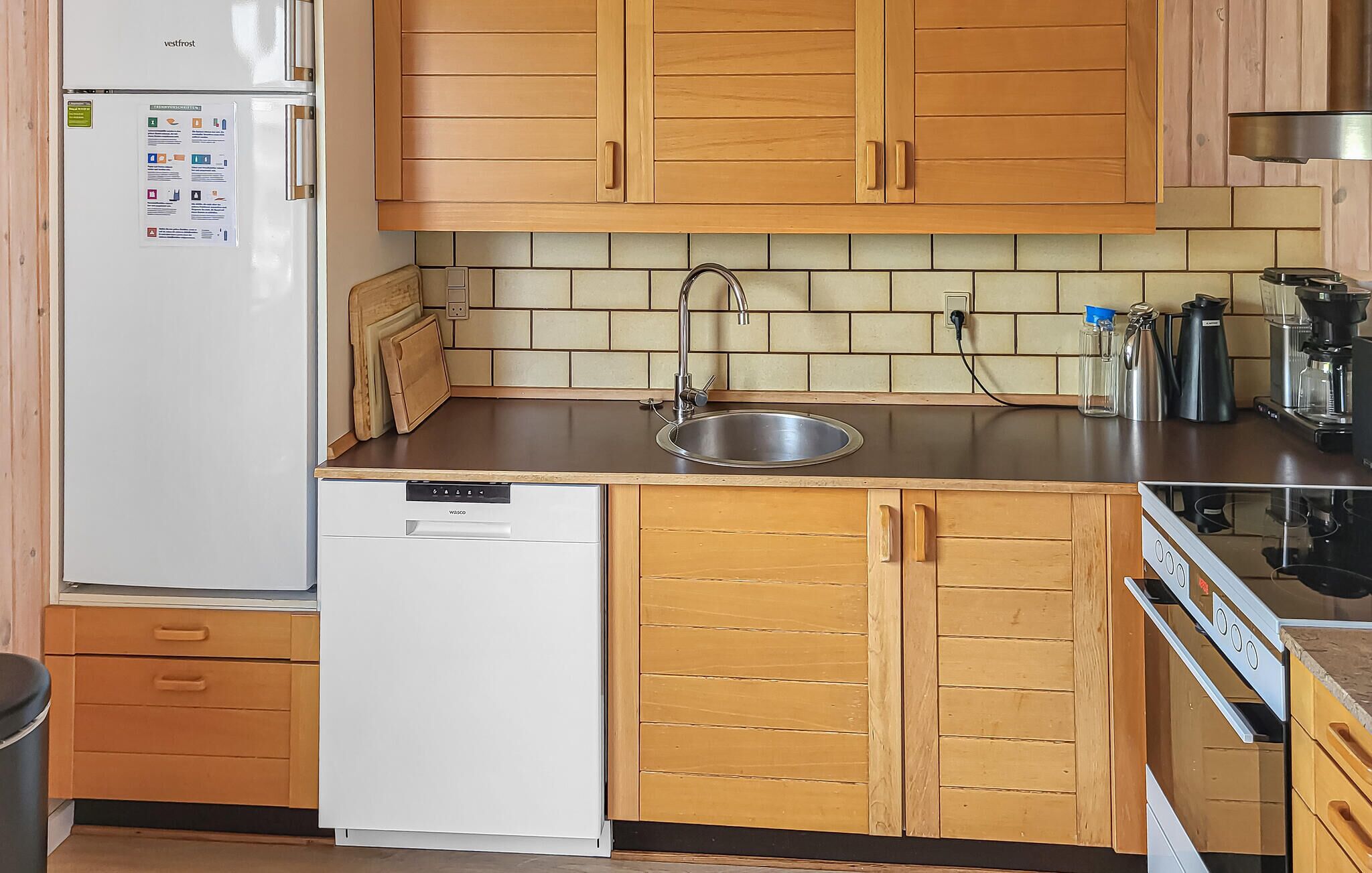 A compact kitchen with wooden cabinets, a stainless steel sink, a white dishwasher, and a coffee maker.