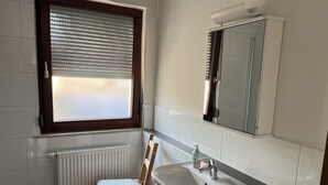 A bathroom with a window, a radiator, a wooden chair, a sink, and a mirror. - Brigittes Fremdenzimmer (Leonberg)