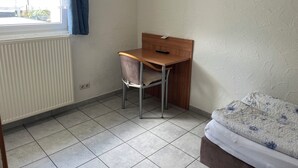 A small room with a wooden desk, a chair, a bed with a floral bedspread, a radiator, and a window with curtains. - Brigittes Fremdenzimmer (Leonberg)
