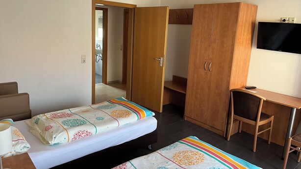 A hotel room with two beds, a desk, a wardrobe, and a TV. - Brigittes Fremdenzimmer (Leonberg)