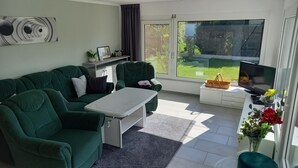 Ein Wohnzimmer mit einem grünen Sofa, einem weißen Couchtisch, einem Fernseher und einem Fenster mit Blick auf Grünflächen.