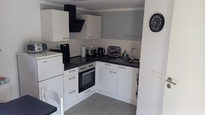 Una cocina compacta con armarios blancos, una encimera negra, horno empotrado, cafetera y un reloj de pared.
