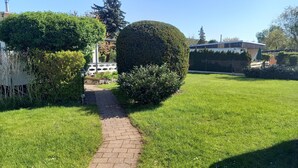Ein gepflasterter Weg führt durch eine gepflegte Grünfläche mit ordentlich geschnittenen Büschen und einer weißen Struktur im Hintergrund.