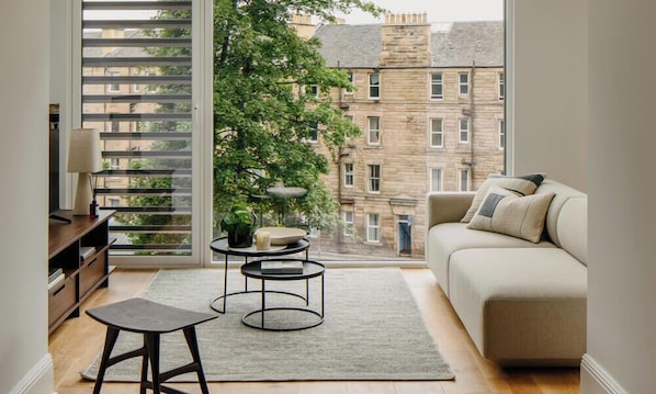 A living room with a sofa, two round tables, a small stool, and a view of buildings through a large window.